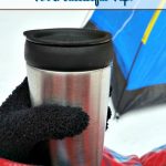 Snow on ground with tent in the background and a hand with a black glove holding a stainless steel mug and covered in red plaid blanket with a text overlay that says "Winter Camping Tips For A Successful Trip".